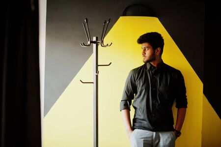 Confident young indian man in black shirt standing near yellow wall.