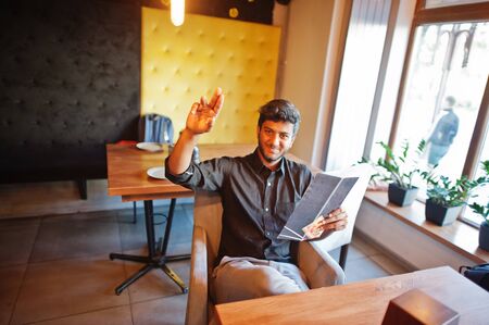 Confident young indian man in black shirt sitting at cafe, read menu and calling waiter.の写真素材