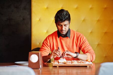 Confident young indian man in orange sweater sitting at pizzeria and eat pizza.の写真素材