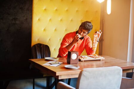 Confident young indian man in orange sweater sitting at pizzeria and eat pizza.の写真素材