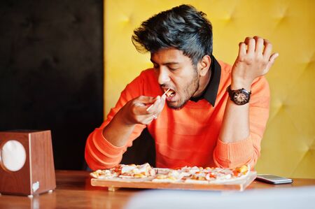 Confident young indian man in orange sweater sitting at pizzeria and eat pizza.の写真素材