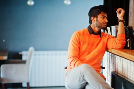 Confident young indian man in orange sweater sitting bar counter at cafe.の写真素材