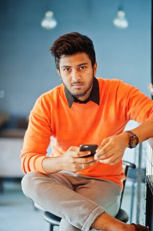 Confident young indian man in orange sweater sitting bar counter at cafe with mobile phone at hand.