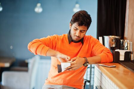 Confident young indian man in orange sweater sitting bar counter at cafe and calculated with the...