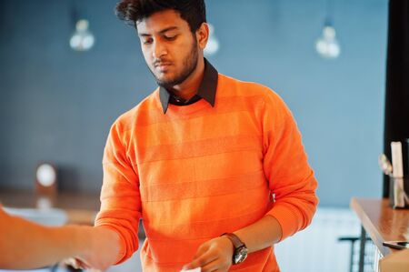 Confident young indian man in orange sweater sitting bar counter at cafe and calculated with the...