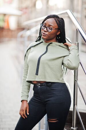 City portrait of positive young dark skinned female wearing green hoody and eyeglasses.