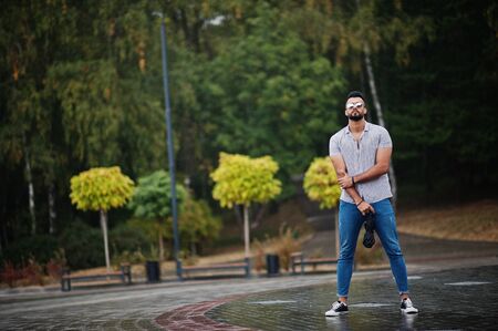 Fashionable tall arab beard man wear on shirt, jeans and sunglasses with umbrella posed at rain on park square.の写真素材