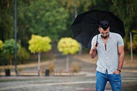 Fashionable tall arab beard man wear on shirt, jeans and sunglasses with umbrella posed at rain on park square.の写真素材