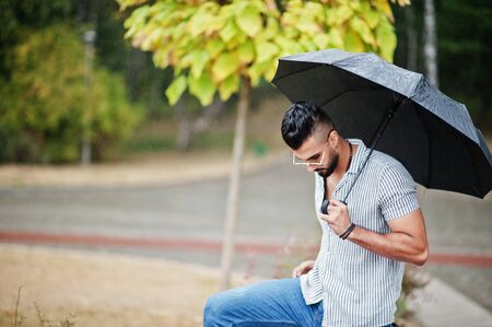Fashionable tall arab beard man wear on shirt, jeans and sunglasses with umbrella posed at rain on park square.の写真素材