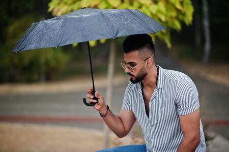 Fashionable tall arab beard man wear on shirt, jeans and sunglasses with umbrella posed at rain on park square.の写真素材