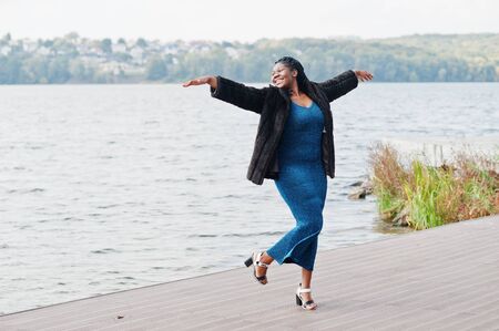 African american dark skinned plus size model posed in a blue shiny dress and black fur coat against...