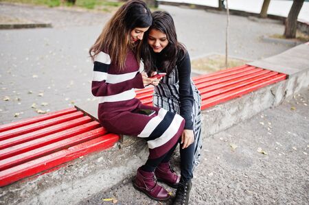 Portrait of two young beautiful indian or south asian teenage girls in dress sitting on bench and...