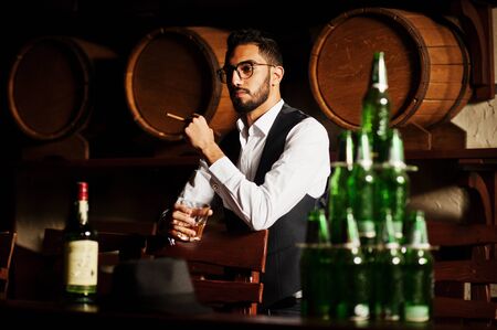 Handsome well-dressed arabian man with glass of whiskey and cigar posed at pub.の写真素材