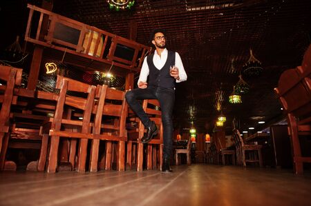 Handsome well-dressed arabian man with glass of whiskey and cigar posed at pub.