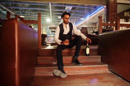 Handsome well-dressed arabian man with glass of whiskey and cigar posed at pub.