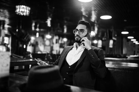 Handsome well-dressed arabian man with glass of whiskey and cigar hold mobile phone,  posed at pub.