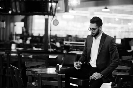 Handsome well-dressed arabian man with glass of whiskey and cigar posed at pub.