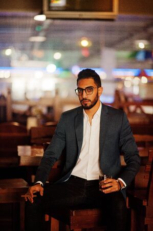Handsome well-dressed arabian man with glass of whiskey and cigar posed at pub.