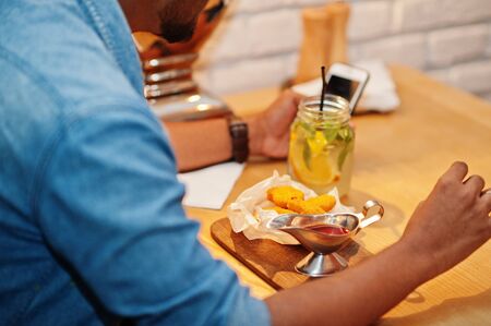 Portrait of handsome successful bearded south asian, young indian freelancer in blue jeans shirt sitting in cafe with chicken nuggets and lemonade. Hold mobile phone at hand.の写真素材