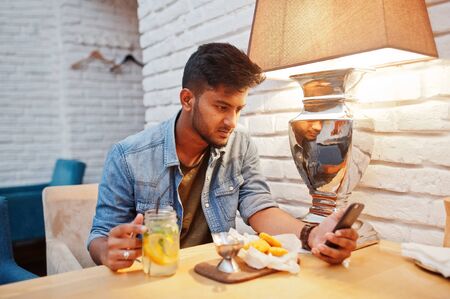 Portrait of handsome successful bearded south asian, young indian freelancer in blue jeans shirt sitting in cafe with chicken nuggets and lemonade. Hold mobile phone at hand.の写真素材
