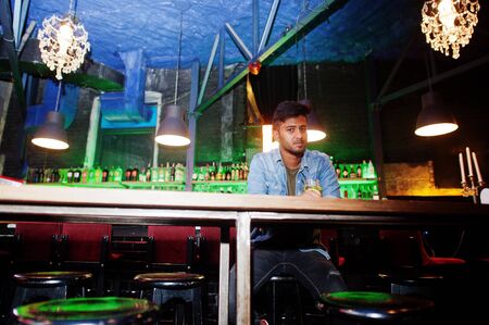 Portrait of handsome successful bearded south asian, young indian freelancer in blue jeans shirt sitting in night club against bar counter with cocktail and having a rest.の写真素材