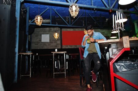 Portrait of handsome successful bearded south asian, young indian freelancer in blue jeans shirt sitting in night club against bar counter with cocktail and having a rest.の写真素材