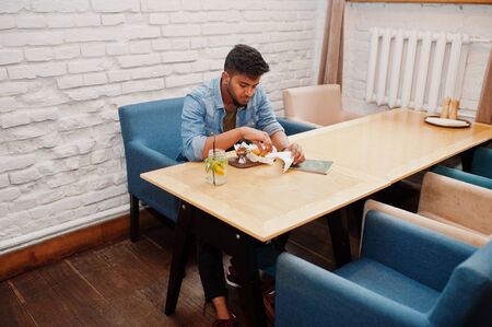 Portrait of handsome successful bearded south asian, young indian freelancer in blue jeans shirt sitting in cafe with chicken nuggets and lemonade.の写真素材