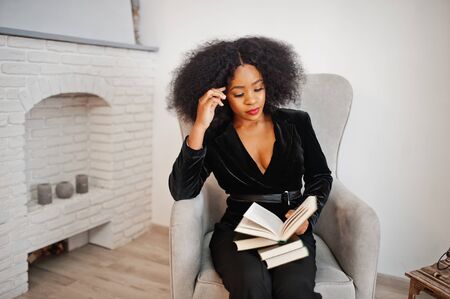 Stylish african american woman in black posed at room against fire place read books.の写真素材