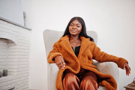 Stylish african american woman in orange coat posed at room sitting at chair.の写真素材