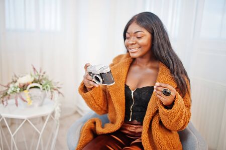 Stylish african american woman in orange coat posed at room sitting at chair