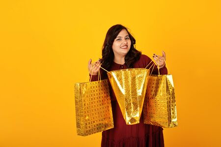 Attractive south asian woman in deep red gown dress posed at studio on yellow background with golden shopping bags.の写真素材