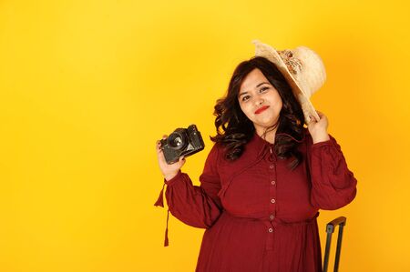 Attractive south asian traveler woman in deep red gown dress, hat posed at studio on yellow background with suitcase amd old vintage photo camera.の写真素材