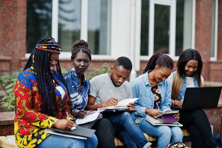 Group of five african college students spending time together on campus at university yard. Black afro friends studying at bench with school items, laptops notebooks.の写真素材