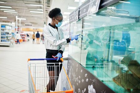 African woman wearing disposable medical mask and gloves shopping in supermarket during coronavirus pandemia outbreak. Black female chooses fish at epidemic time.の写真素材