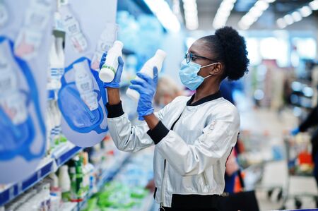 African woman wearing disposable medical mask and gloves shopping in supermarket during coronavirus pandemia outbreak. Black female choose milk at epidemic time.の写真素材