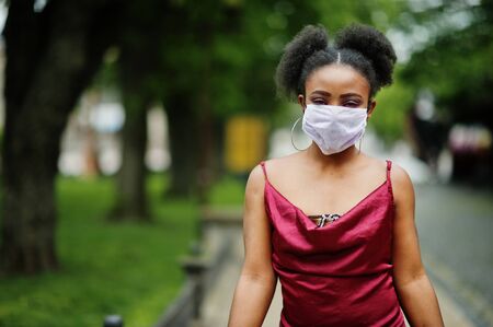 Covid-19, infectious virus. African woman with curly hair, wears red silk dress and medical disposable mask, cares about her health and protects in dangerious situation.の写真素材