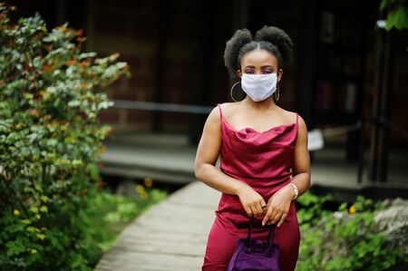 Covid-19, infectious virus. African woman with curly hair, wears red silk dress and medical disposable mask, cares about her health and protects in dangerious situation.の写真素材