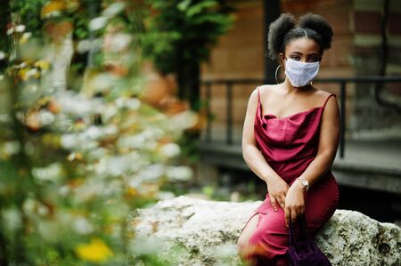 Covid-19, infectious virus. African woman with curly hair, wears red silk dress and medical disposable mask, cares about her health and protects in dangerious situation.の写真素材