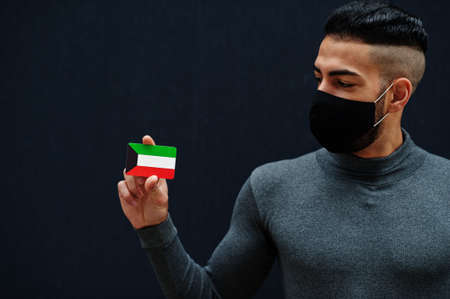 Middle eastern man in gray turtleneck and black face protect mask show Kuwait flag isolated background.の素材 [FY310157263811]