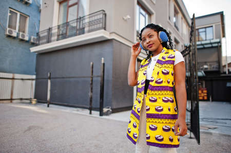 Stylish african american women in yellow jacket posed and earphones on street against modern...