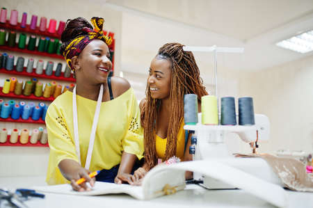Two african dressmaker woman sews clothes on sewing machine and looking at designer album in tailor...