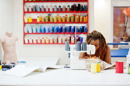 Young african dressmaker woman sews clothes on sewing machine at tailor office. Black seamstress women.の写真素材