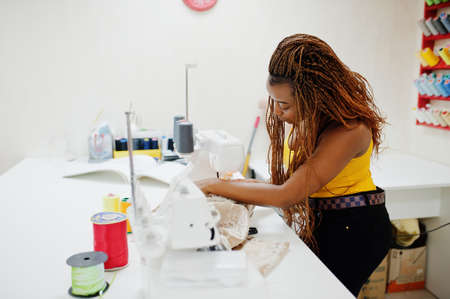 Young african dressmaker woman sews clothes on sewing machine at tailor office. Black seamstress women.の写真素材