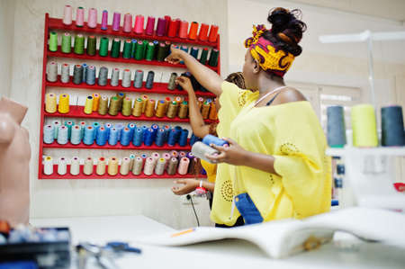 Two african dressmaker woman sews clothes on sewing machine and selects threads at tailor office....