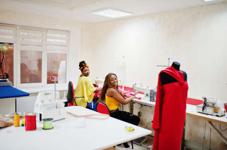 Two african dressmaker woman sews clothes on sewing machine at tailor office. Black seamstress girls.の写真素材