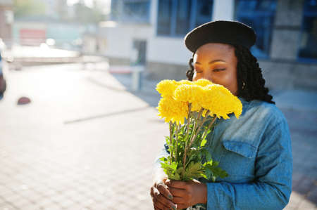 Stylish fashionable african american women in jeans wear and black beret with yellow flowers bouquet posed outdoor in sunny day against blue modern building.の写真素材