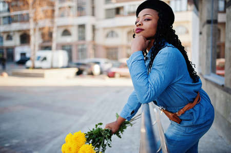 Stylish fashionable african american women in jeans wear and black beret with yellow flowers bouquet posed outdoor in sunny day against blue modern building.の写真素材