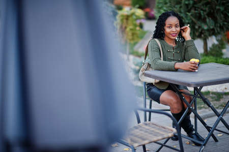 Stylish fashionable african american women in green sweater and black skirt posed outdoor cafe, sitting by table with cup of coffee.の写真素材
