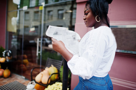 Stylish african american women in white blouse and blue jeans posed near cafe with newspaper.の写真素材