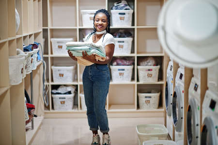 Cheerful african american woman with towels in hands near washing machine in the self-service...